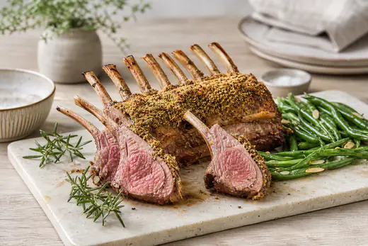 Un carré d’agneau rôti, croûte dorée aux herbes, coupé en côtes sur une planche, avec du romarin et des haricots verts