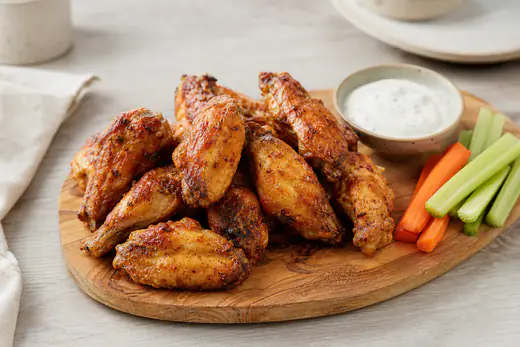Ailes de poulet à la peau dorée et croustillante, empilées sur une planche en bois avec un bol de sauce et des bâtonnets de céleri