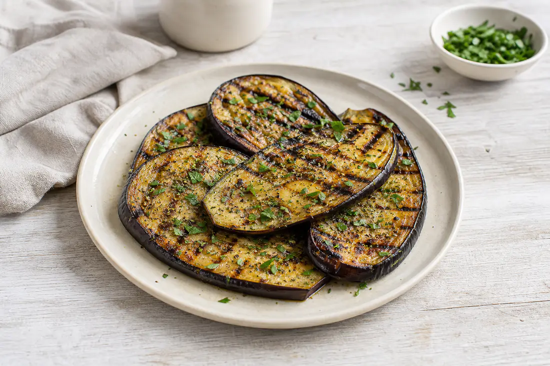 Tranches d’aubergine grillées, dorées et marquées, parsemées d’origan et de persil, posées sur une assiette