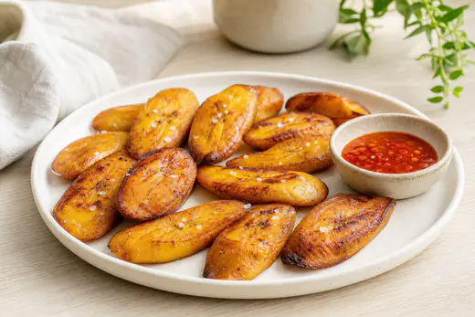Rondelles de banane plantain mûre dorées et caramélisées en un seul lit sur une assiette blanche, avec une pincée de sel et un petit bol de sauce pimentée