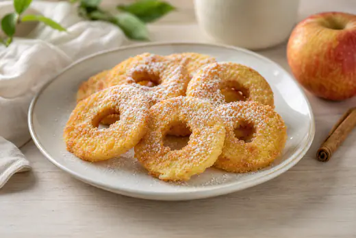 Des rondelles de pomme panées et dorées, avec le trou central, saupoudrées de sucre glace sur une assiette, avec une pomme et de la cannelle