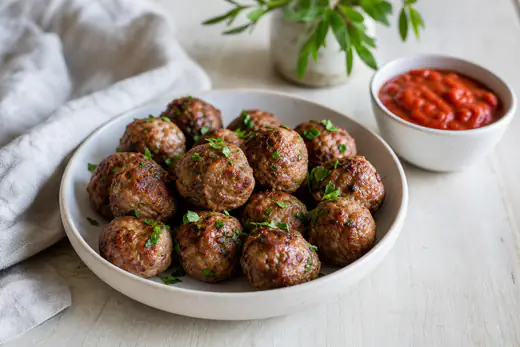 Des boulettes de viande rondes et dorées dans un plat avec du persil haché, à côté d’un bol de sauce tomate