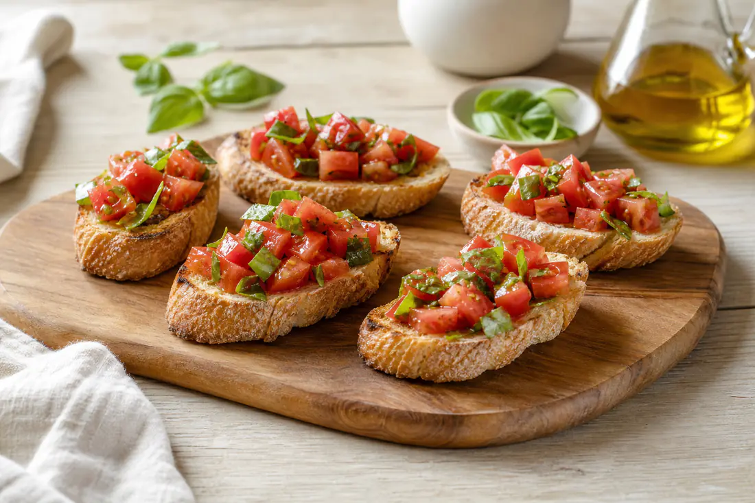 Six tranches de pain grillé garnies de dés de tomates fraîches, de feuilles de basilic et d’huile d’olive, servies sur une planche en bois