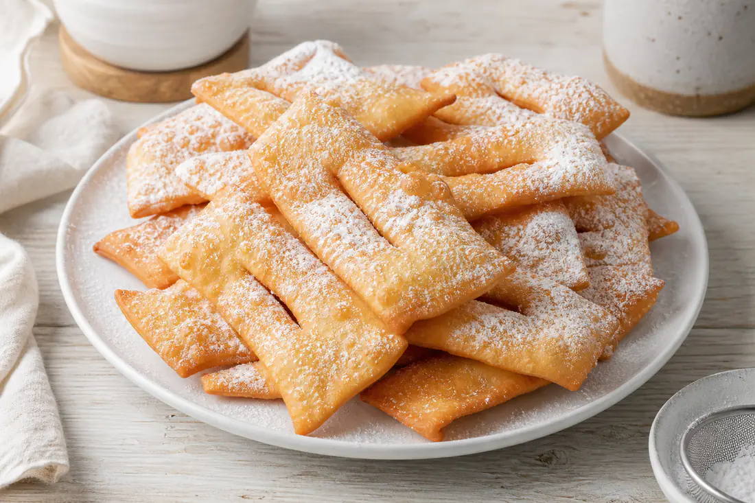 Bugnes lyonnaises en rectangles fendus, dorées et saupoudrées de sucre glace, empilées sur une assiette blanche