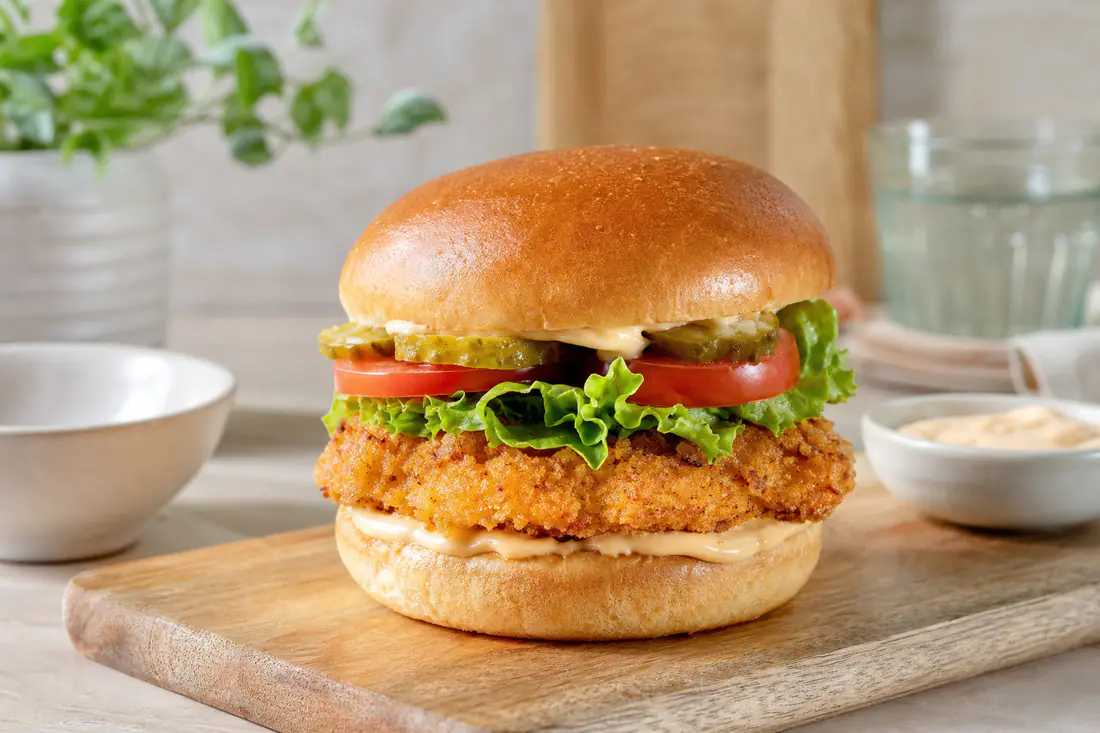 Burger de poulet pané dans un pain à burger toasté, avec salade, tomate, cornichons et sauce, posé sur une planche en bois