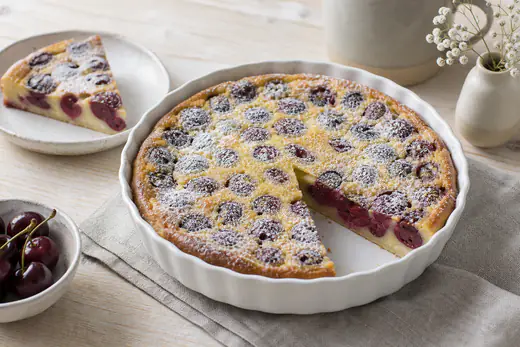 Clafoutis rond doré dans un plat à gratin de 18 cm, cerises rouges visibles à la surface, sucre glace tamisé, une part prélevée