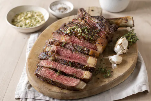 Côte de bœuf avec l’os, croûte brune bien saisie, tranchée en lamelles roses à cœur sur une planche avec du beurre à l’ail et du thym