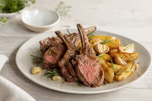 Quatre côtelettes d’agneau avec l’os, bien dorées, posées sur une assiette avec du romarin, de l’ail et des pommes de terre rôties