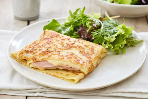 Crêpes salées pliées en carré, garnies de jambon et d’emmental gratiné doré, servies sur une assiette avec de la salade