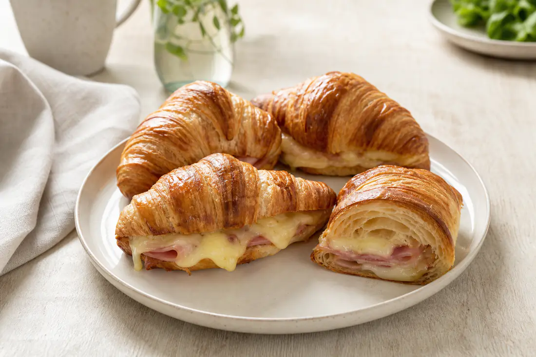 Quatre croissants au jambon dorés, l’un coupé en deux pour montrer le jambon, la béchamel et l’emmental fondu, servis sur une assiette
