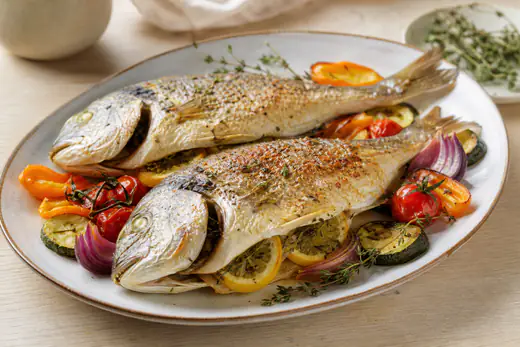 Deux dorades entières grillées à la peau dorée, farcies de citron et de thym, servies sur une assiette avec des légumes rôtis