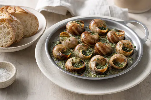 Une douzaine d’escargots au beurre persillé qui grésillent dans un plat à escargots, avec du pain de campagne