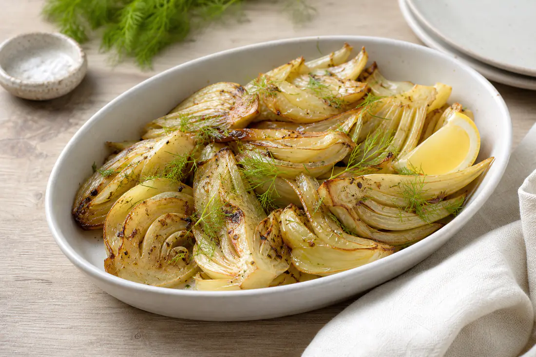 Quartiers de fenouil rôtis, caramélisés et dorés sur les bords, avec du citron et des herbes fraîches, dans un plat blanc