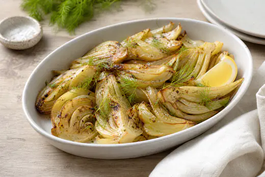 Quartiers de fenouil rôtis, caramélisés et dorés sur les bords, avec du citron et des herbes fraîches, dans un plat blanc