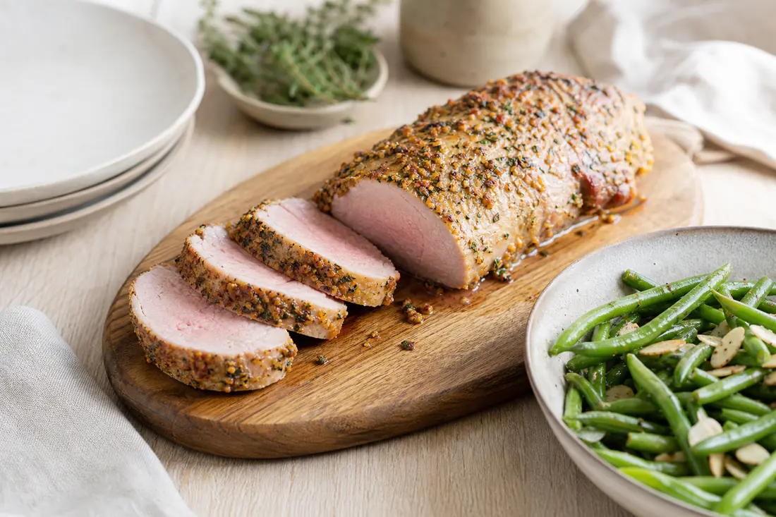 Filet mignon de porc entier doré à la croûte de moutarde et d’herbes, tranché en rondelles roses sur une planche en bois avec des haricots verts