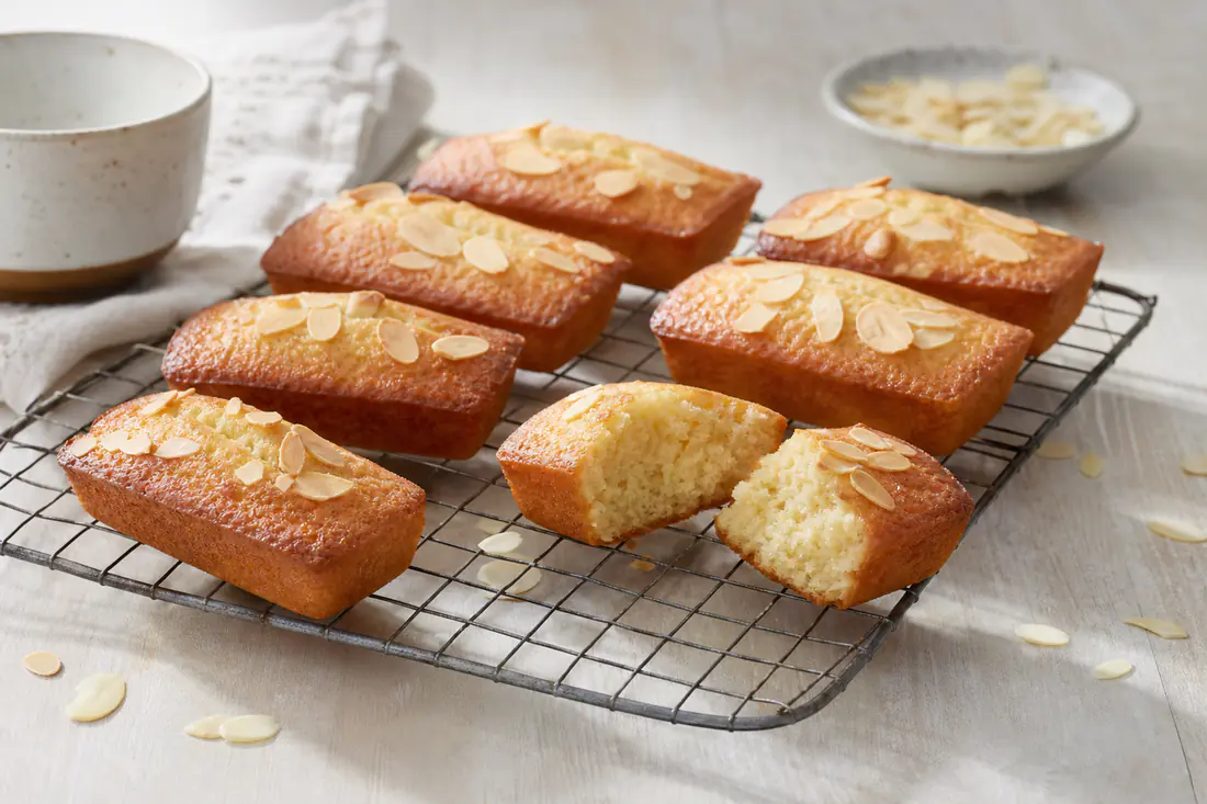 Huit financiers rectangulaires dorés aux amandes posés sur une grille, un financier coupé en deux montrant une mie moelleuse