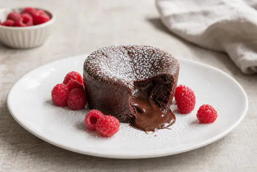 Fondant au chocolat démoulé sur une assiette, entamé pour montrer le cœur de chocolat coulant, avec des framboises et du sucre glace