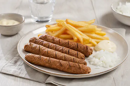 Quatre fricadelles allongées et dorées, entaillées en biais, servies avec des frites, de la mayonnaise et de l’oignon cru