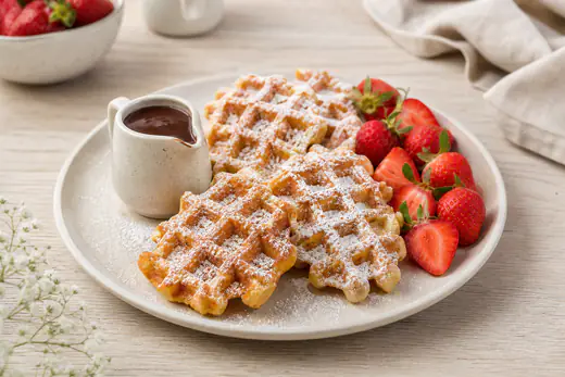 Plusieurs petites gaufres dorées à l’alvéole marquée, saupoudrées de sucre glace, avec des fraises et un petit pot de chocolat fondu