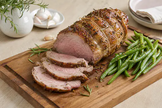 Un gigot d’agneau désossé et ficelé, doré, tranché en rondelles rosées sur une planche, avec du romarin, de l’ail et des haricots verts