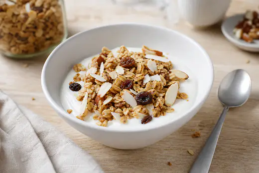 Granola maison doré avec flocons d’avoine, amandes et raisins secs, dans un bol blanc avec du yaourt nature
