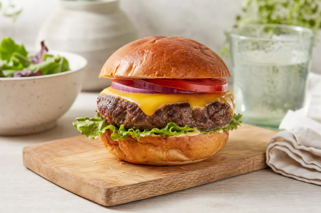 Hamburger maison avec un steak haché épais, cheddar fondu, salade, tomate et oignon rouge dans un pain grillé, posé sur une planche en bois