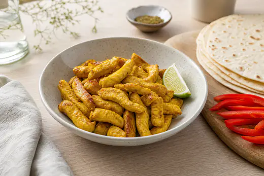 Lanières de poulet dorées et épicées dans un bol, avec des quartiers de citron vert, des poivrons et des tortillas à côté