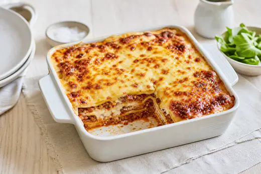Petite lasagne bolognaise gratinée dans un plat carré, fromage doré, avec une part découpée laissant voir les couches de pâtes et de viande