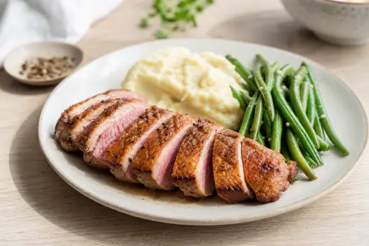 Magret de canard rosé tranché en lamelles, avec une peau dorée et croustillante, servi avec de la purée et des haricots verts