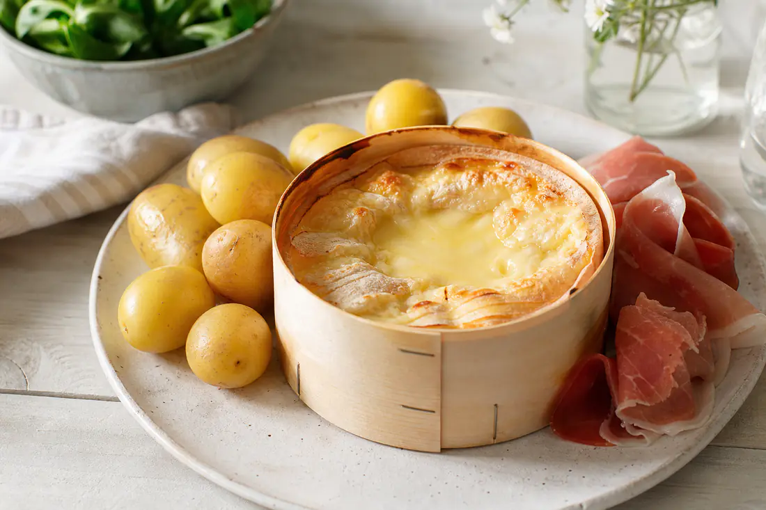Un mont d’or chaud dans sa boîte en bois, la croûte dorée et le fromage crémeux, entouré de pommes de terre grenaille et de charcuterie