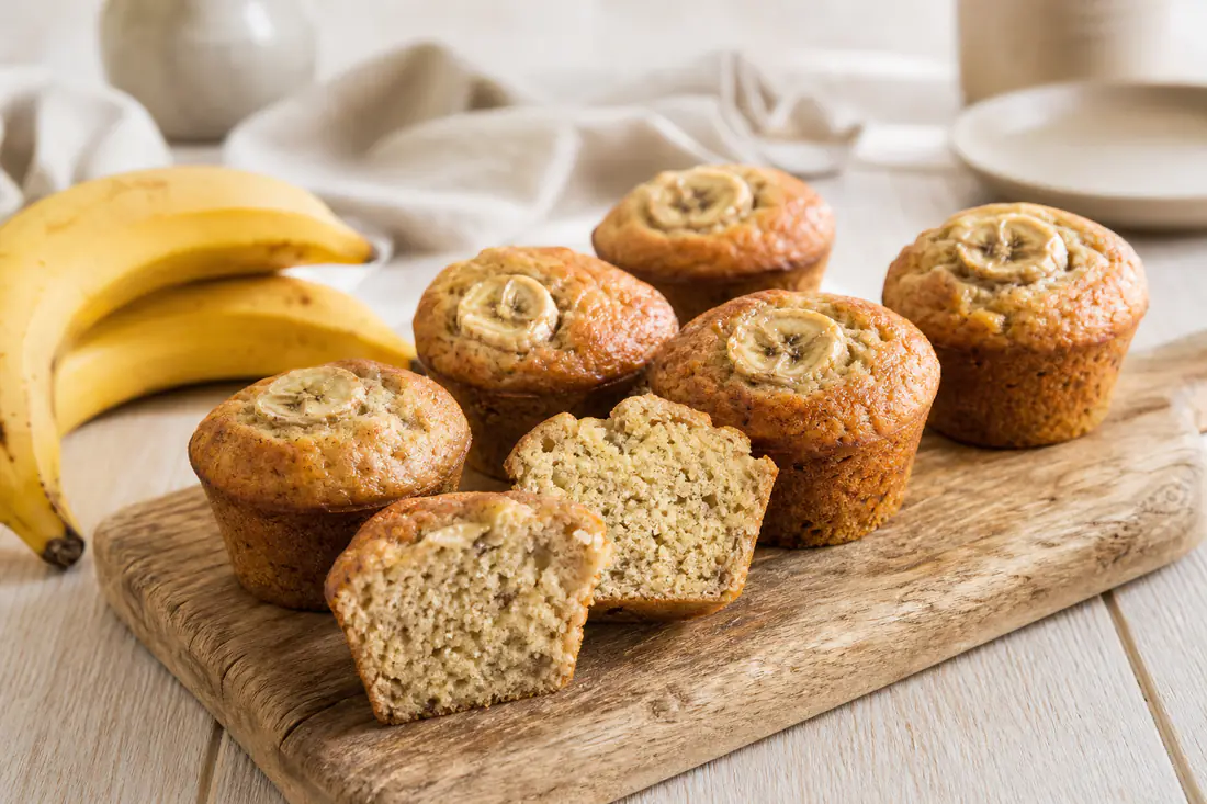 Muffins à la banane dorés avec des rondelles de banane sur le dessus, l’un coupé en deux, servis sur une planche en bois avec des bananes mûres
