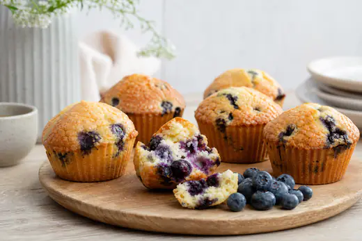 Muffins aux myrtilles bien dorés et bombés, l’un coupé en deux avec des myrtilles bleues visibles dans la mie, avec une poignée de myrtilles fraîches à côté