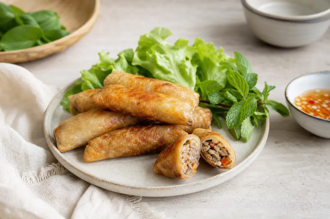 Plusieurs nems dorés et croustillants dont un coupé en deux, avec feuilles de salade, menthe et un bol de sauce nuoc-mâm