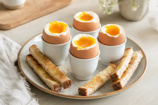 Quatre œufs à la coque dans des coquetiers, le dessus ouvert sur un jaune coulant, avec des mouillettes de pain grillées et du beurre