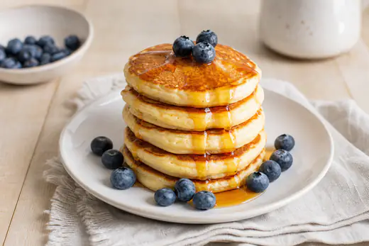 Une pile de petits pancakes ronds et épais, dorés, nappés de sirop d’érable et accompagnés de myrtilles fraîches sur une assiette