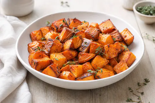 Morceaux de patate douce rôtis, dorés et caramélisés sur les bords, avec quelques brins de thym dans un plat blanc