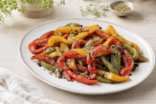 Lamelles de poivrons rouges, jaunes et verts grillées, aux bords légèrement noircis, dans une assiette avec des herbes