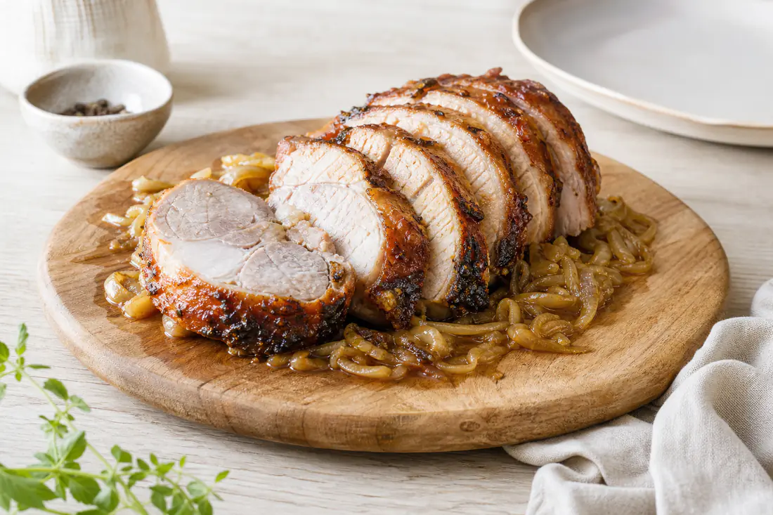 Rouelle de porc épaisse rôtie, croûte dorée et brillante, posée sur des oignons caramélisés et tranchée en parts sur une planche