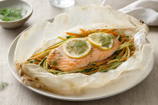 Papillote de papier cuisson ouverte avec un pavé de saumon rosé, des rondelles de citron, de la julienne de carottes et de courgettes et de l’aneth