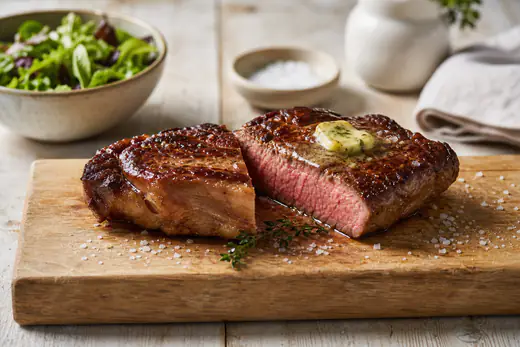 Un pavé de bœuf épais, grillé, avec une croûte brune, coupé en deux pour montrer le cœur rosé, posé sur une planche avec du sel et du thym