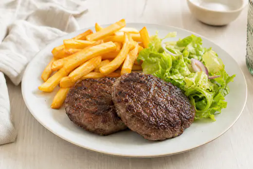 Deux steaks hachés bien dorés posés sur une assiette, avec des frites et une salade verte