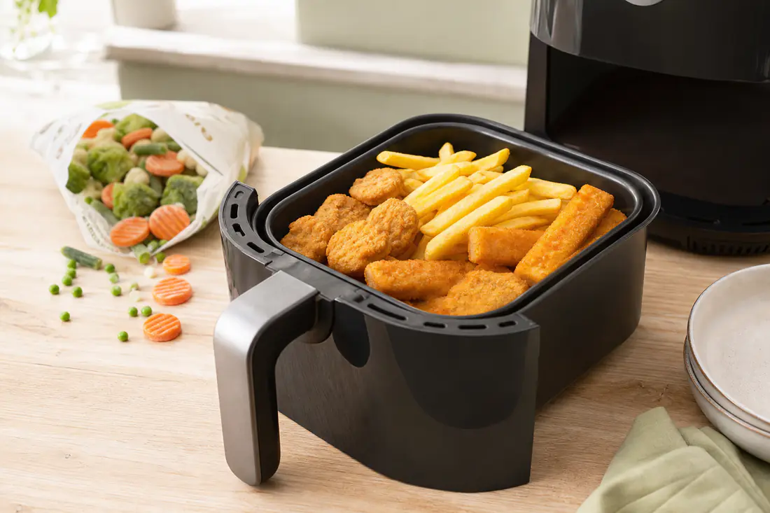 Panier d’airfryer garni de frites, nuggets de poulet et bâtonnets de poisson dorés, avec un sachet de légumes surgelés à côté