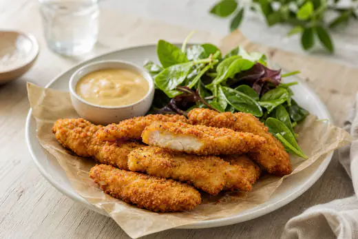 Des tenders de poulet panés et dorés posés sur du papier, avec un bol de sauce et une salade verte