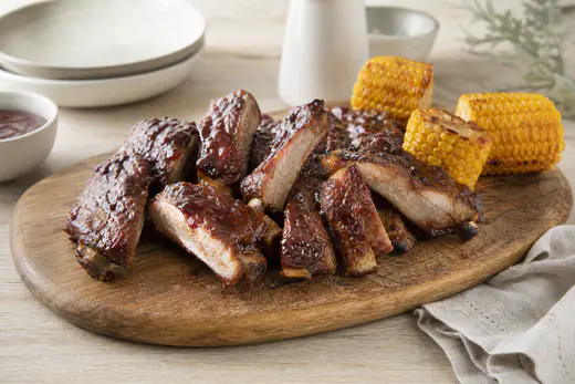 Travers de porc laqués à la sauce barbecue, brillants et caramélisés, coupés en portions avec les os visibles, servis sur une planche avec du maïs