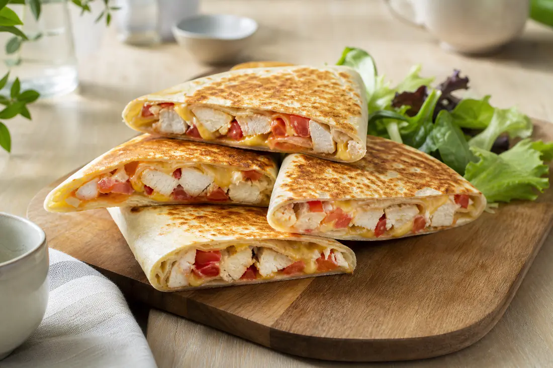 Quatre wraps grillés au poulet et au fromage, coupés en biais pour montrer la garniture, posés sur une planche avec de la salade et des tomates